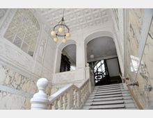 Treppenhaus in einem historischen Gebäude mit weißem Marmor, verzierten Geländern und Wandreliefs. Oben befinden sich Arkadenbögen, eine kunstvolle Deckenverzierung und ein Kronleuchter mit runden Glühbirnen.