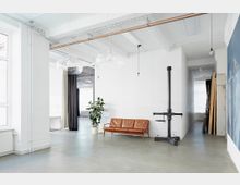 Minimalistisch eingerichteter, heller Raum mit weißen Wänden, einer Ledercouch, einer Zimmerpflanze und großen Glaslampen an der Decke. Im Hintergrund sind ein Durchgang mit Vorhängen und diverse Arbeitsgeräte an einer Wand zu sehen.