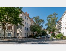 Straßenszene in einem Wohnviertel mit mehrstöckigen, hellen Mietshäusern und Balkonen. Im Vordergrund stehen Bäume, geparkte Fahrzeuge und ein Fahrrad, während Personen die Straße überqueren; Verkehrszeichen und eine ruhige Atmosphäre prägen das Bild.