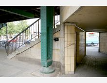 Straßenunterführung mit gelb gefliesten Wänden, einer grünen, genieteten Stahlstütze und einer Betontreppe, die nach oben führt. Im Hintergrund sind Fahrräder, ein Gehweg und Geschäfte zu sehen.
