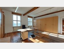 Heller Büroraum mit Holzboden, weißen Wänden und freiliegenden Holzbalken an der Decke. Der Raum enthält einen Schreibtisch mit Bürostuhl, eine Fensterfront mit Sprossenfenstern sowie eine Wand mit hohen Holzschränken.