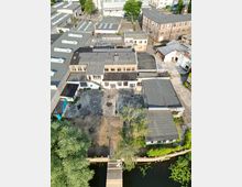 Luftaufnahme eines Industriegeländes mit mehreren flachen, teils verfallenen Gebäuden im Backstein- und Betondesign. Im Vordergrund befinden sich ein kleiner Kanal mit Seerosen, eine Holzbrücke sowie ein Baum und ein unbefestigter Hof.