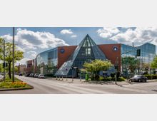 Großes, modernes Gebäude mit einer markanten Glaspyramide in der Mitte, flankiert von rotbraunen Backsteinwänden und Glasfassaden. Der Eingangsbereich ist von Bäumen und Begrünung umgeben, und eine Straße mit Autos und Fußwegen verläuft im Vordergrund.