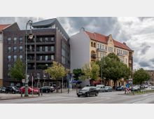 Straßenszene mit zwei nebeneinanderliegenden Wohngebäuden: links ein modernes Gebäude aus dunklem Backstein mit Balkonen, rechts ein gelb gestrichenes historisches Haus mit verzierten Fassaden und roten Dachziegeln. Bäume und parkende Autos säumen den Straßenrand, im Vordergrund fahren Autos auf einer mehrspurigen Straße.