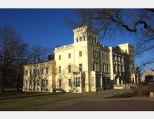 Gelbes neoklassizistisches Gebäude mit einem Turm und symmetrischen Fenstern, umgeben von einem Park mit kahlen Bäumen unter klarem blauem Himmel. Im Vordergrund befindet sich eine gepflasterte Fläche und Rasen.