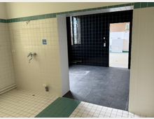 Gefliester Innenraum mit weißen Wandfliesen und grünem Rand, links mit einem Wandwasserhahn und Leitungen. Im Durchgang liegt ein dunkler, quadratisch gefliester Bereich mit schwarzer Rückwand und Fenster, der zu einem weiteren hell gefliesten Nebenraum führt.