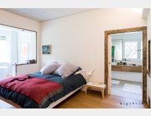 Ein modernes Schlafzimmer mit einem Doppelbett, bedeckt von einer dunkelblauen Decke und einem roten Überwurf. Rechts befindet sich eine Schiebetür, die ein Badezimmer mit weißer Einrichtung und großen Fenstern zeigt; links ist ein großer Fensterbereich mit weißen Vorhängen und minimalistischem Dekor sichtbar.