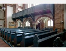 Innenraum einer Kirche mit Holzbänken in dunklem Blau, gotischen Backsteinbögen und einer Empore mit verzierter Brüstung. Im Hintergrund ist eine kleine Orgel sowie Fenster mit Bleiverglasung sichtbar.