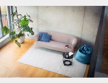 Heller Wohnbereich mit Betonwand und großen Fensterflächen, ausgestattet mit einem hellen Sofa auf einem großen hellen Teppich. Daneben stehen eine große Zimmerpflanze, ein kleiner runder Beistelltisch und ein blauer Sitzsack auf Holzboden.