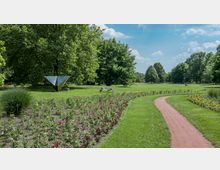 Grüne Parklandschaft mit einer geschwungenen roten Backsteinspur, umgeben von Blumenbeeten und gepflegtem Rasen. Im Hintergrund stehen große Bäume, und eine moderne Metallskulptur ist auf der Wiese zu sehen.