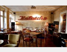 Ein gemütlicher Raum mit einer Mischung aus Stühlen und Tischen aus verschiedenen Epochen, einer neonbeleuchteten Schrift "Kollasche" an der Wand und einem alten Klavier in der Ecke. Die Wände sind verputzt, aber ungestrichen, was dem Raum ein rustikales Aussehen verleiht; Lampen und Tageslicht durch Fenster sorgen für Beleuchtung.