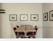 Minimalistisch eingerichteter Raum mit einem rechteckigen Holztisch, umgeben von vier roten Stühlen. An der weißen Wand hängen fünf eingerahmte Schwarz-Weiß-Fotografien in gleichmäßiger Anordnung.