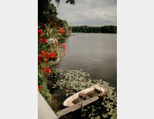Blick über einen ruhigen See mit Seerosen am Ufer; im Vordergrund liegt ein kleines Ruderboot an einem einfachen Holzsteg. Am gegenüberliegenden Ufer verläuft eine dichte Baumlinie unter bewölktem Himmel, links rahmen unscharfe rote Blüten das Bild.