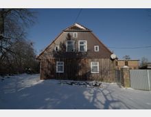 Ein freistehendes, älteres Wohnhaus mit steilem Satteldach und Holzfassade steht in einer verschneiten Hof- oder Gartenfläche. Mehrere kleine Fenster, eine Satellitenschüssel an der rechten Seite sowie angrenzende Zäune und Nebengebäude sind vor blauem Winterhimmel zu sehen.
