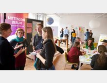 Eine helle, offene Workshop-Umgebung mit mehreren Gruppen von Personen, die an Tischen sitzen oder in kleinen Gruppen stehen und sich austauschen. Im Hintergrund sind Tafeln mit Notizzetteln, eine Leinwand mit der Aufschrift „Mittendrin Berlin!“ sowie ein hängender Globus zu sehen.