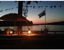 Eine Terrasse mit Holztisch und Beleuchtung durch Laternen im Vordergrund, umgeben von Blumenkübeln. Im Hintergrund ein See bei Sonnenuntergang, mit einer aufgezogenen Fahne und sanften Hügeln am Horizont.