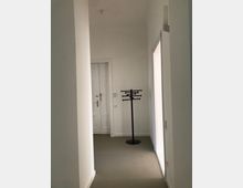 Ein schlichter, heller Flur mit weißen Wänden und einer beigefarbenen Bodenfläche. Im Hintergrund steht ein schwarzer Kleiderständer vor einer zweiflügeligen weißen Tür.