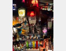 Eine Bar mit schummeriger Beleuchtung, roten Vorhängen und einem Retro-Ambiente. Im Vordergrund sind Cocktails, bunte Flüssigkeiten in Reagenzgläsern und ein Absinthspender zu sehen, während im Hintergrund Regale mit zahlreichen Spirituosenflaschen und eine beleuchtete Theke erkennbar sind.