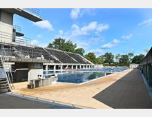 Freibad mit mehreren Becken, darunter ein Sprungbecken mit Sprungturm und ein großes Schwimmbecken, umgeben von erhöhten Sitztribünen aus Beton. Im Hintergrund sind Bäume und blauer Himmel sichtbar.
