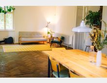 Heller Wohnraum mit Holzboden, einer grauen Couch und gelbem gestreiftem Teppich. Im Vordergrund steht ein Holztisch mit Stühlen, und im Hintergrund sind ein Sessel, Pflanzen sowie eine Stehlampe zu sehen.