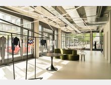 Ein heller und moderner Showroom mit großen Glasfassaden, durch die Tageslicht einfällt. Im Raum befinden sich Kleiderständer mit Kleidung, eine grüne modulare Sitzbank, ein schwarzer runder Tisch und ein Bereich mit Tischen und Stühlen im hinteren Teil.