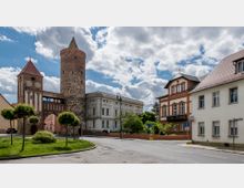 Stadtszene mit einem historischen Stadttor aus Fachwerk und Backstein, flankiert von einem mittelalterlichen Rundturm. Im Hintergrund stehen ein klassizistisches weißes Gebäude und ein rotes Ziegelhaus, umgeben von Bäumen und einer Straße mit gepflastertem Gehweg.