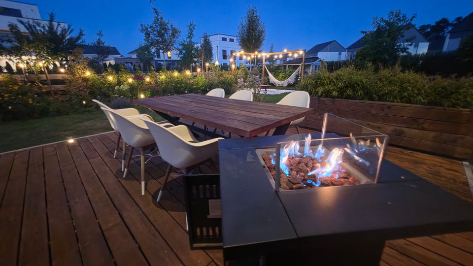 Holzterrasse in einem Garten bei Dämmerung mit großem Esstisch und mehreren weißen Stühlen sowie einem modernen Gas-Feuertisch im Vordergrund. Im Hintergrund sind bepflanzte Beete, Lichterketten, eine Hängematte unter einer Pergola und Wohnhäuser zu sehen.
