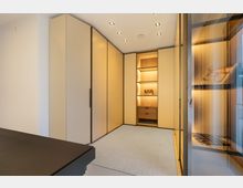 Moderner begehbarer Kleiderschrank mit deckenhohen, cremefarbenen Einbauschränken und einem beleuchteten Holzregal mit Schublade in der Mitte. Rechts sind gläserne Regaleinsätze mit integrierter Beleuchtung und schwarze Rahmen zu sehen, darüber eingelassene Decken-Spots.