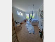 Helles Kinderzimmer mit Holzfußboden, einem Einzelbett, einem Schreibtisch mit Stuhl und einem Hängesitz in der Mitte des Raumes. Vor den großen Fenstern mit Blick ins Grüne liegt ein geometrisch gemusterter Teppich, an den Wänden sind Dekorationen und Zeichnungen befestigt.