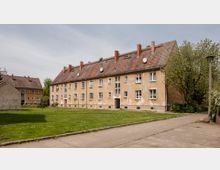 Mehrfamilienhaus mit zwei Stockwerken und einem Satteldach, umgeben von einer Rasenfläche und einigen Bäumen. Die gelblich verputzte Fassade zeigt Abnutzungsspuren, und auf dem Dach sind mehrere Satellitenschüsseln angebracht.