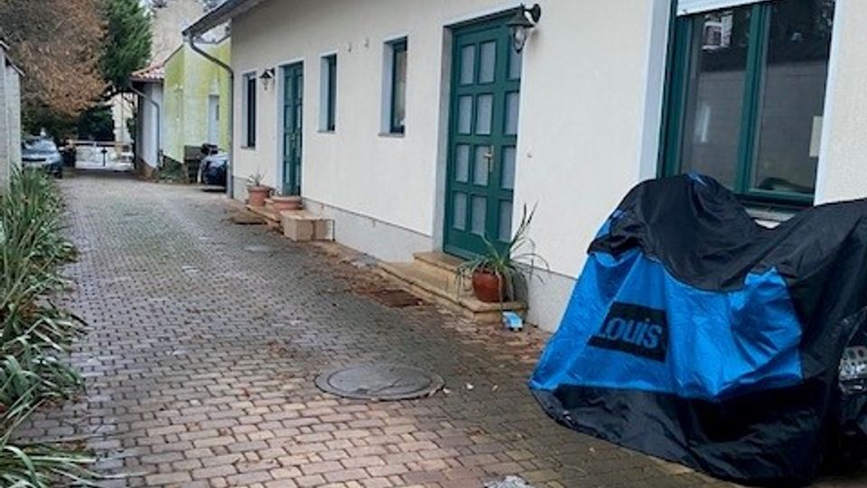 Schmaler, gepflasterter Hofweg entlang eines weißen Wohnhauses mit grün gerahmten Fenstern und grüner Haustür sowie Satteldach. Am Rand stehen Pflanzen und ein abgedecktes Fahrzeug; der Weg führt zu weiteren Gebäuden und Bäumen im Hintergrund.