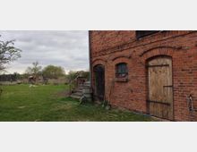 Eine Ziegelsteinfassade eines ländlichen Gebäudes mit Holz- und Metalltüren sowie einem kleinen Fenster mit Sprossen. Im Vordergrund befindet sich eine grüne Wiese, während im Hintergrund Bäume, ein Trampolin und landwirtschaftliche Gegenstände sichtbar sind.