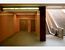 Untergrundbereich mit orangefarbenen Fliesenwänden, einem Aufzug in der Mitte, einer Tür daneben und auf der rechten Seite einer Rolltreppe sowie einer Treppe. Oben befindet sich ein beleuchtetes Schild mit der Aufschrift „Aufzug“.