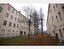 Innenhof mit herbstlich gefärbtem Laubboden, umgeben von beige-farbenen, verputzten Altbauten mit sichtbaren Abnutzungsspuren an den Fassaden. Mehrere kahle Bäume durchziehen den Hof, der von schmalen Wegen und vereinzelten Sitzbänken gesäumt ist.