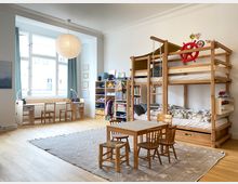 Helles Kinderzimmer mit hohen Decken und großem Fenster, einem langen Schreibtisch mit zwei Stühlen sowie Regal- und Stauraummöbeln an der Wand. Rechts steht ein großes Holz-Hochbett mit Leiter und Seilen; davor ein kleiner Tisch mit vier Stühlen auf einem Teppich.