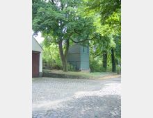 Kopfsteinpflasterstraße mit einem kleinen, grünen, holzverkleideten Turmgebäude unter hohen Bäumen. Im Vordergrund ist ein gepflasterter Platz, und im Hintergrund sind weitere Bäume und Vegetation sichtbar.