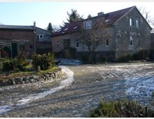 Gehöft mit alten Gebäuden, darunter ein Wohnhaus mit rotem Ziegeldach und verputzter Fassade sowie ein angrenzendes Wirtschaftsgebäude. Im Vordergrund ein gepflasterter Hof mit vereinzeltem Eis und kleinen, bewachsenen Beeten.