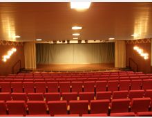 Ein kleiner Veranstaltungssaal mit roten Polstersitzen, einer erhöhten Bühne mit geschlossenen Vorhängen und Holzwänden mit moderner Beleuchtung. Die Decke ist mit einer hellen Paneelstruktur versehen, und der Raum ist gleichmäßig beleuchtet.