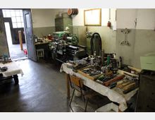 Werkstatt mit Maschinenwerkbank und Werkzeugen. Der Raum zeigt eine Drehmaschine an der Wand, mehrere Werkzeuge und Metallteile auf einem Arbeitstisch sowie karge Wände und einen offenen Zugang zu einem angrenzenden Bereich mit Tageslicht.