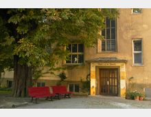 Fassade eines alten Gebäudes mit bräunlicher Wandfarbe, großen Fenstern und einem Eingang mit der Aufschrift „Theater auf der Zitadelle“. Vor dem Gebäude stehen unter einem großen Baum zwei rote Sitzbänke auf gepflastertem Boden.