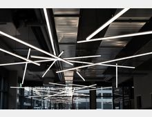 Moderne Innenansicht eines Gebäudes mit auffälliger Deckenbeleuchtung aus langen, kreuzförmig angeordneten LED-Leuchten. Im Hintergrund sind große Fenster und ein urbaner Außenbereich mit Bäumen und Gebäuden sichtbar.