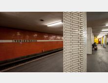 U-Bahn-Station mit oranger Kachelwand und der Aufschrift „Yorckstraße“, weißen gefliesten Säulen, einer wartenden Person auf einer Bank und gelben Wandbereichen im Hintergrund.