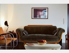 A cozy living room with a green upholstered vintage sofa flanked by wooden armrests, a small side table with a modern lamp, and orange chairs with chrome frames. A framed photograph is mounted on a white wall above the sofa, and a bowl of fruit sits on a white coffee table in the foreground.