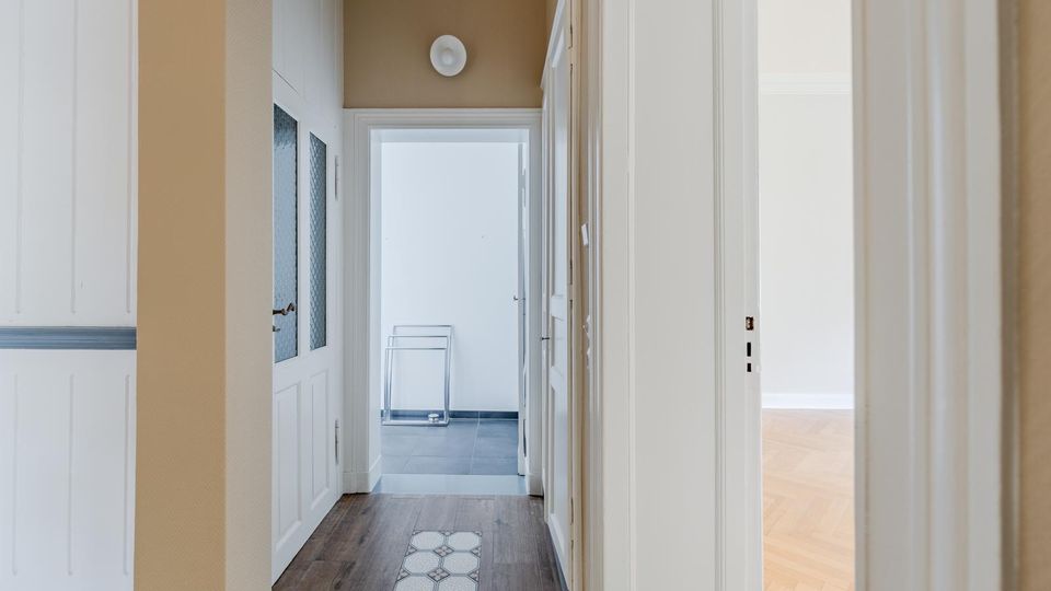 Schmaler Wohnungsflur mit beigefarbenen Wänden, weißen Türen und Holzfußboden mit dekorativem Fliesenstreifen in der Mitte. Am Ende führt eine Tür in einen hellen Raum mit grauen Bodenfliesen, seitlich öffnet sich ein weiterer Raum mit Parkettboden.