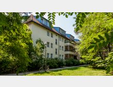 Mehrstöckiges Wohngebäude mit hellem Putz und roten Ziegeldächern, umgeben von dichtem Grün und Bäumen. Ein Fahrrad steht neben einem Fußweg, der an einer Straßenlaterne und einem Straßenschild vorbeiführt.