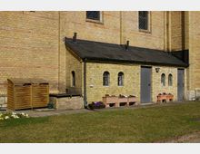 Gelber Backsteinbau mit kleinen, bogenförmigen Fenstern und einem schwarzen Schrägdach, flankiert von Metalltüren. Im Vordergrund stehen bunte Blumenkästen und ein Holzschuppen auf einer gepflasterten Fläche, umgeben von einer Rasenfläche.