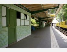 Überdachter U-Bahnhof mit grün gekachelten Wänden, einer geschlossenen Tür und Sitzbänken. Rechts im Bild steht ein gelber U-Bahn-Zug auf den Schienen, während der Bahnsteig mit einem taktilen Leitsystem versehen ist.