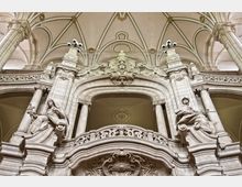 Blick auf ein prächtiges, barockes Innenraumdetail mit hohen dekorativen Säulen, symmetrischen Bögen und kunstvoll geschnitzten Statuen. Die Decke ziert ein Gewölbe mit verzierten Ornamenten und goldenen Akzenten.