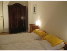 Schlafzimmer mit einem Doppelbett, weißen Bettbezügen und gelben Kissen. Im Hintergrund stehen ein dunkler Holzschrank, ein kleiner Nachttisch mit Lampe sowie ein Bild an der Wand.