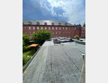 Flachdach mit grauer Bitumenabdichtung, Dachfenstern und niedriger Attika, im Vordergrund mit Sitzkissen. Dahinter ein großer roter Backsteinbau mit vielen weißen Fenstern und Gauben sowie ein begrünter Innenhof unter wolkigem Himmel.